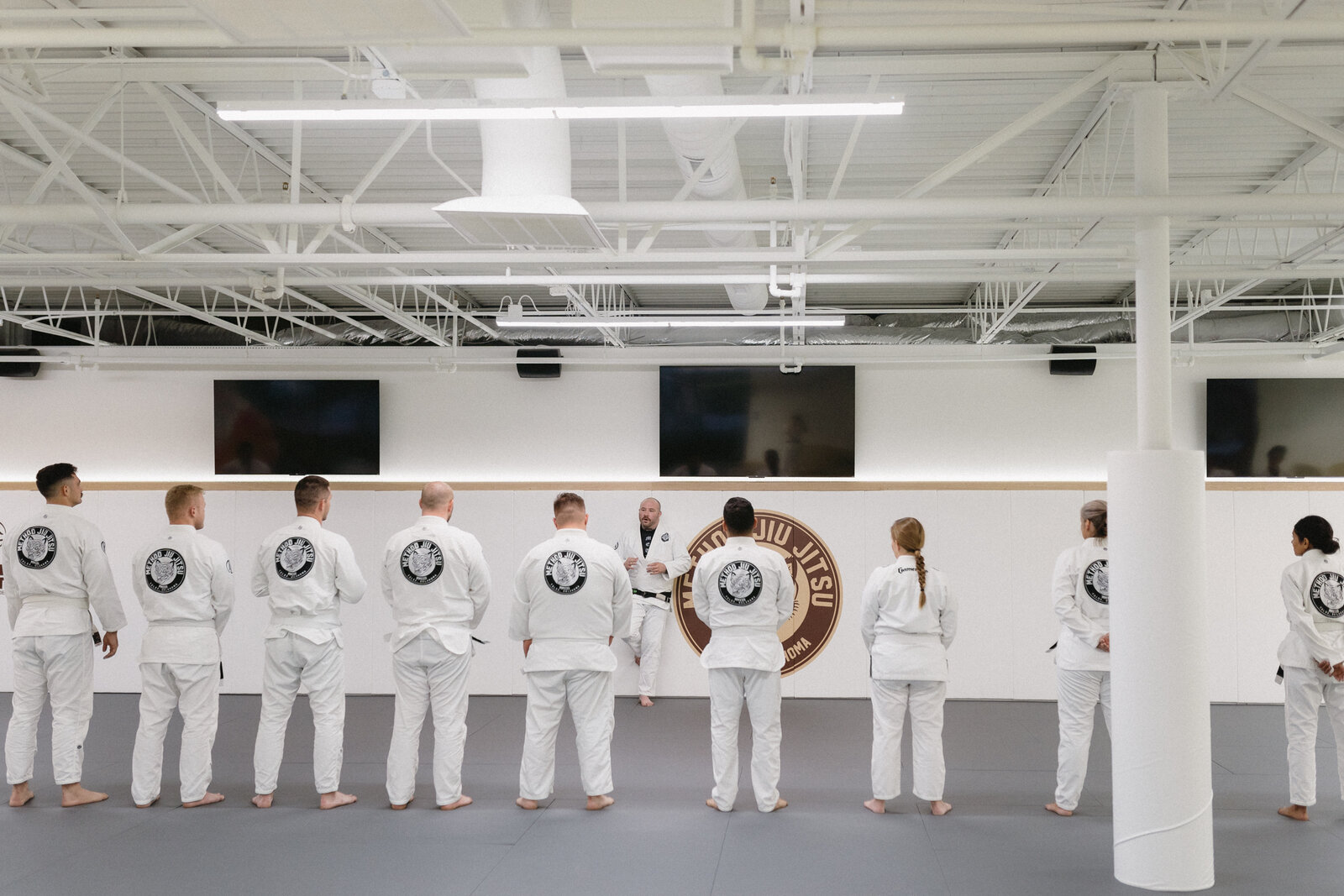 Adult Brazilian jiu-jitsu students in white Method Jiu-Jitsu gis lined up on the mats listening to their instructor during a class in South Tulsa