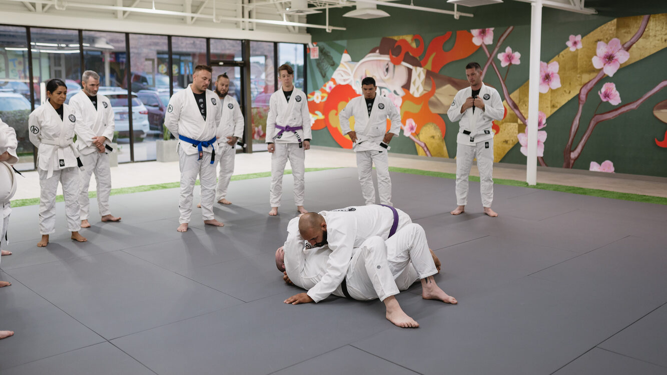 A purple belt pins a black belt's arm beside his head with chest to chest pressure as Method Jiu-Jitsu students watch in South Tulsa