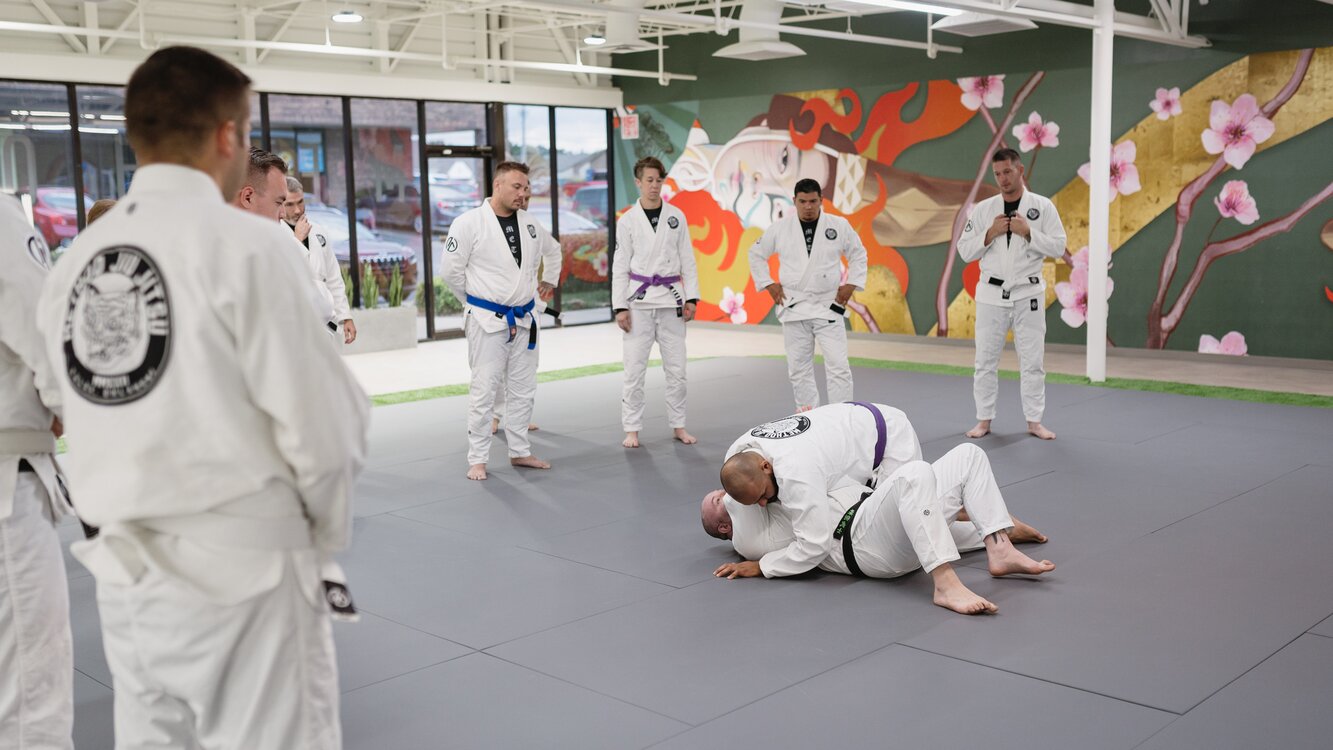 Method Jiu-Jitsu students in white gis watch a black belt pinned on his back under a purple belt's chest-to-chest pressure in South Tulsa