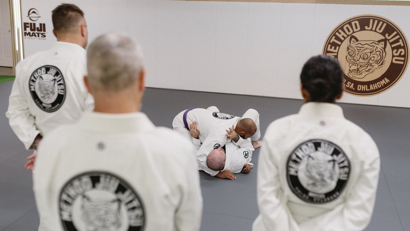 Method Jiu-Jitsu students in white gis watching a purple belt coach demonstrate how to control a partner in the turtle position on the mats in South Tulsa