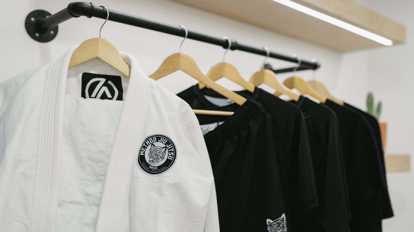 A white Method Jiu-Jitsu gi with a Tulsa, Oklahoma tiger patch on a wide wooden hanger beside black training shorts and shirts on a clothing rack